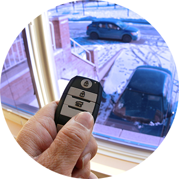 A closeup of a remote starter with someone's finger pressing the button to start it. In the background is a closed, see-through glass window showing the car with snow in the driveway